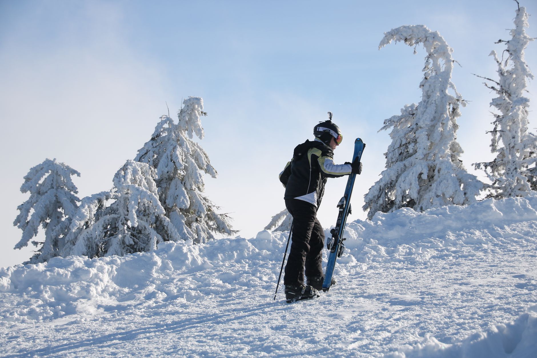 Un skieur solitaire transporte son équipement en montée à travers un paysage enneigé, vêtu d'une tenue de sports d'hiver.
