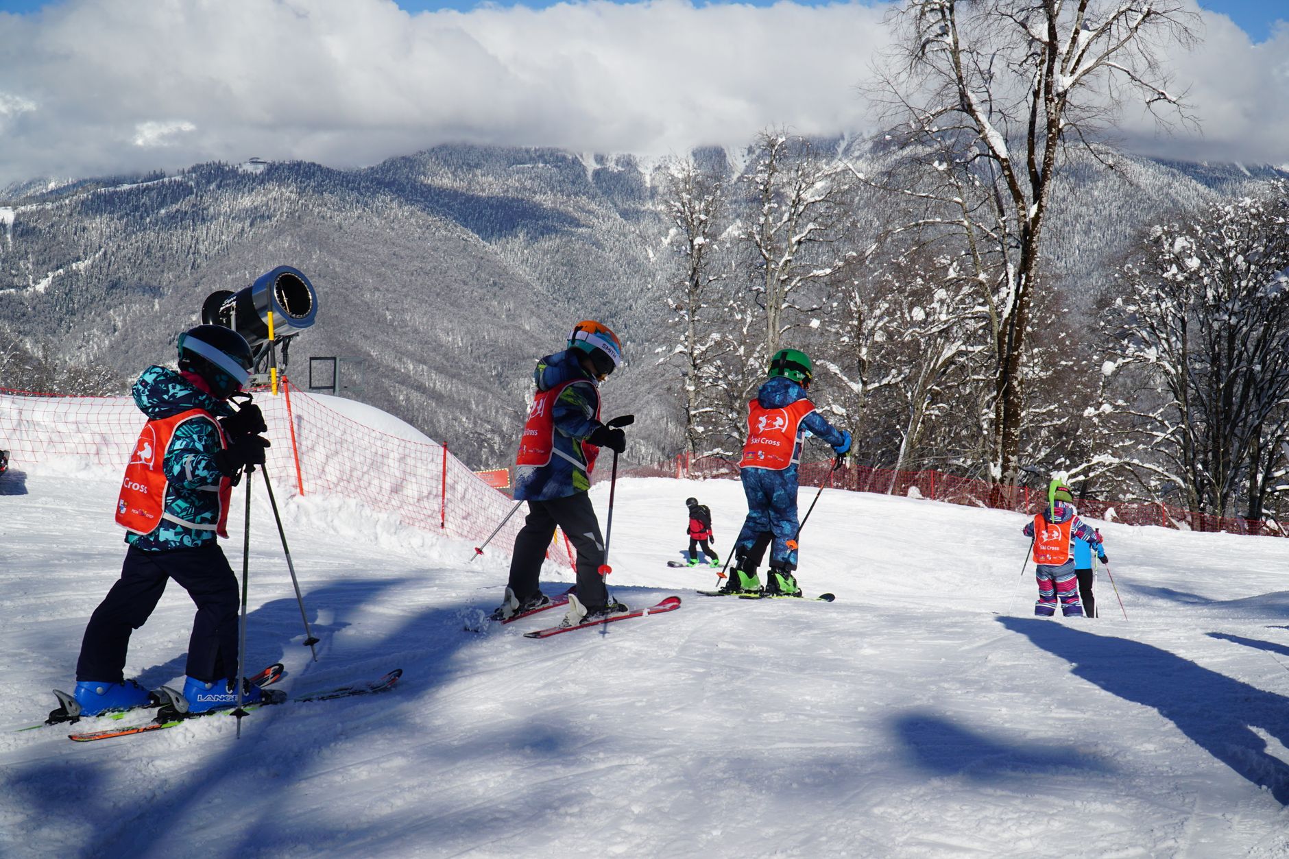 Enfants à ski avec des dossards orange sur une piste enneigée, un canon à neige et une montagne boisée derrière