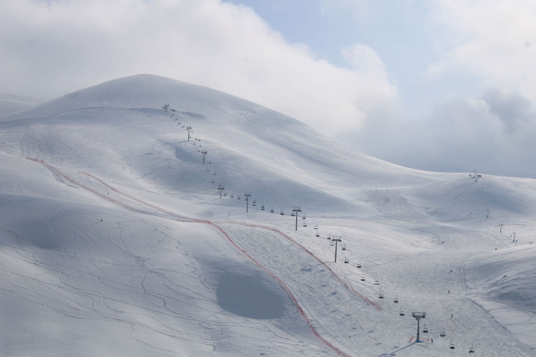 Pistes de ski enneigées avec un télésiège montant vers un sommet arrondi, une piste balisée de filets rouges et des nuages dans le ciel.