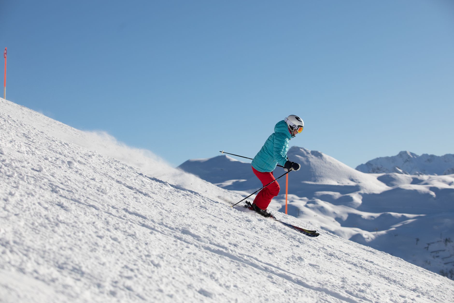 Skieuse en veste turquoise et pantalon rouge descendant une piste enneigée, avec des montagnes couvertes de neige en arrière-plan.