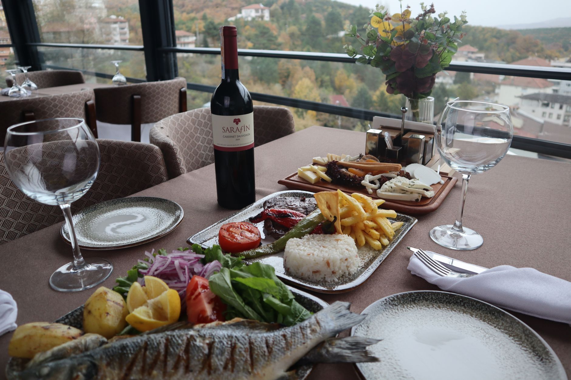 Table de restaurant avec poisson grillé, plateaux de viande, riz et fromages, bouteille de vin et verres, devant une baie vitrée donnant sur des collines boisées.