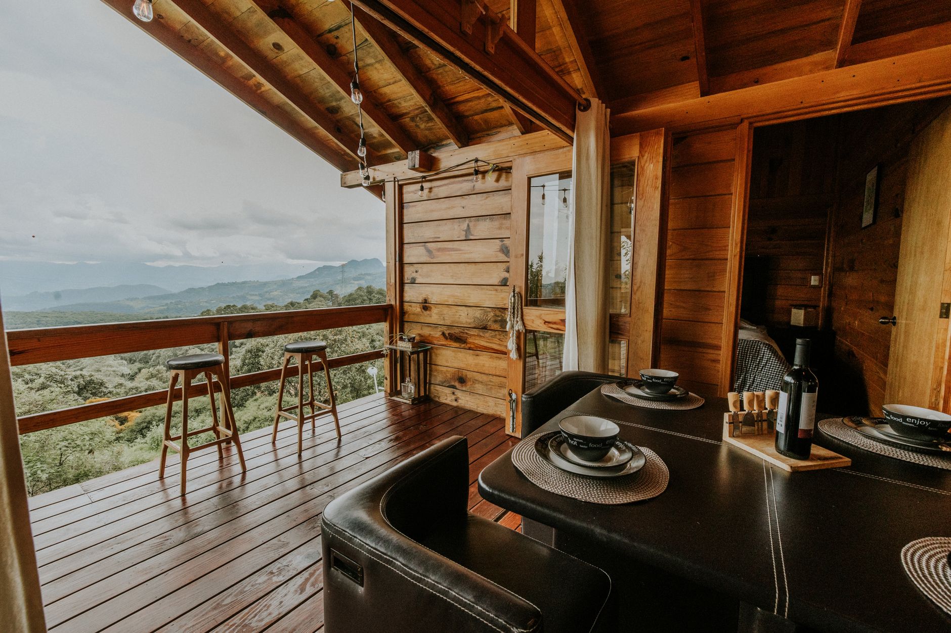 Terrasse en bois d’un chalet avec tabourets et table dressée, face à une vallée boisée et des montagnes sous un ciel nuageux.