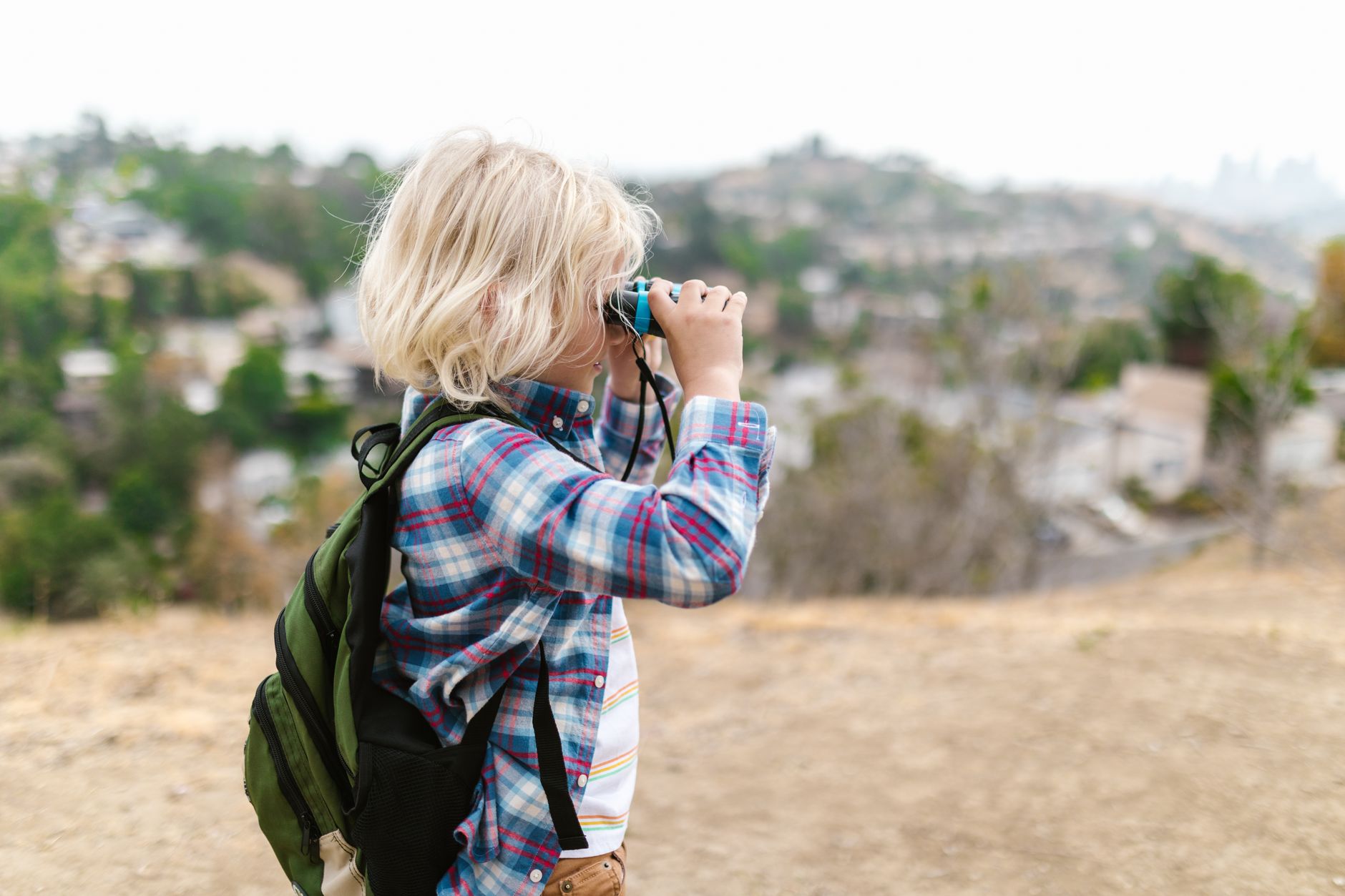 Un jeune enfant portant un sac à dos regarde à travers des jumelles en plein air au sommet d’une colline.